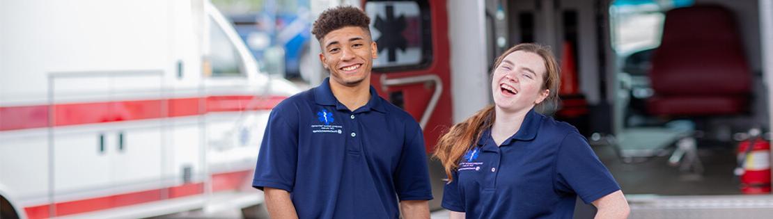 Two paramedic students stand smiling in front of an ambulance